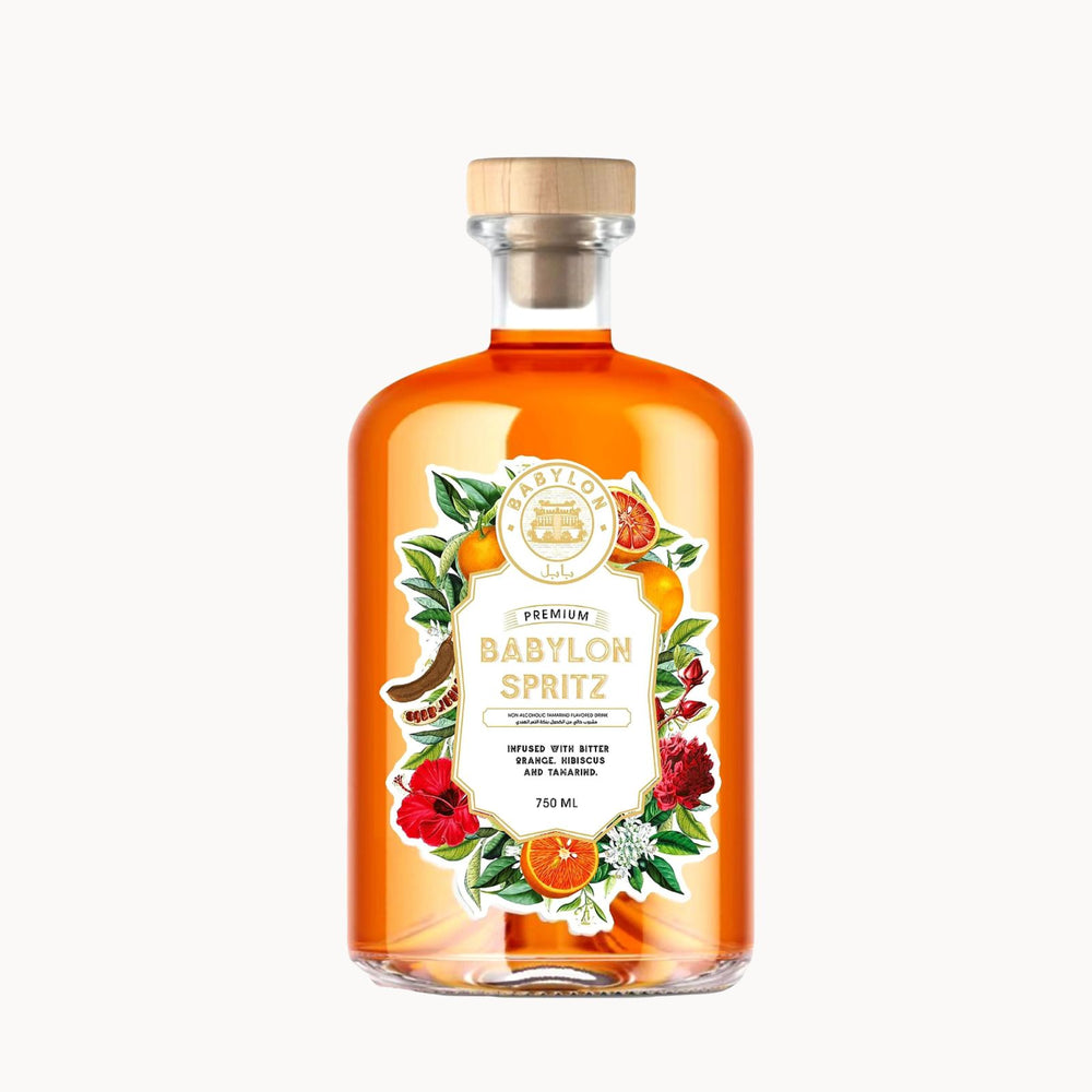 A clear glass bottle of Babylon Spritz with amber-orange liquid, a light wood cap, and a decorative label featuring illustrations of flowers, citrus, and a cocktail glass, shown on a plain white background.
