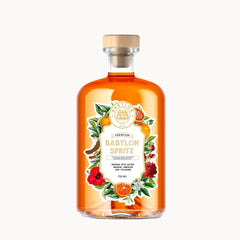 A clear glass bottle of Babylon Spritz with amber-orange liquid, a light wood cap, and a decorative label featuring illustrations of flowers, citrus, and a cocktail glass, shown on a plain white background.
