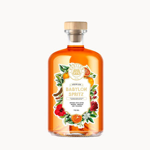 A clear glass bottle of Babylon Spritz with amber-orange liquid, a light wood cap, and a decorative label featuring illustrations of flowers, citrus, and a cocktail glass, shown on a plain white background.