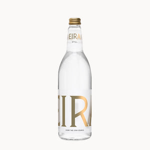 A clear glass bottle of Eira Still water with a gold cap and a white label featuring large gold 'EIR' lettering, set against a plain off-white background.