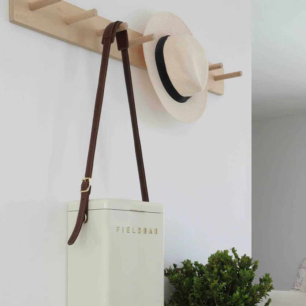 White drinks box with brown strap and 'FIELDBAR' branding hanging on a wooden coat rack against a white wall.