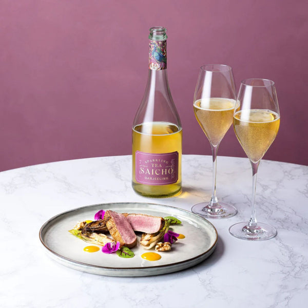 Bottle of wine, two glasses with Darjeeling sparkling tea, and a plate of food on a marble surface with a purple background.