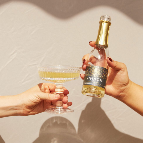 Rienne_Two hands holding a glass of white wine and a bottle labeled 'VINADA' against a neutral background.