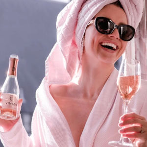Woman in a pink robe with a towel on her head, holding a bottle and glass of pink wine.
