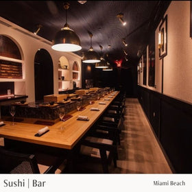 Sushi bar with long wooden counter, chairs, and decor in a dimly lit room.