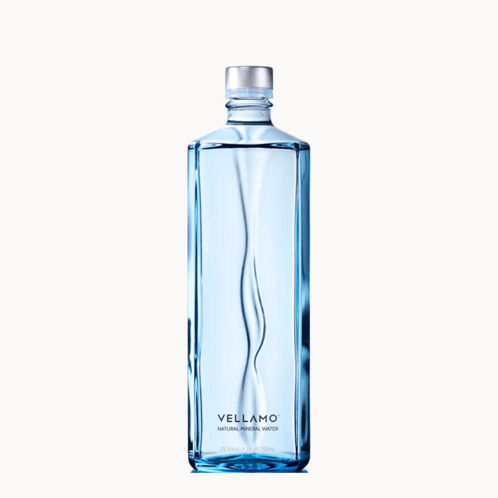 A tall, rectangular glass bottle of Vellamo Natural Mineral Water with a wavy design and a silver cap, centered on a light off-white background.