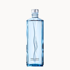 A tall, rectangular glass bottle of Vellamo Natural Mineral Water with a wavy design and a silver cap, centered on a light off-white background.