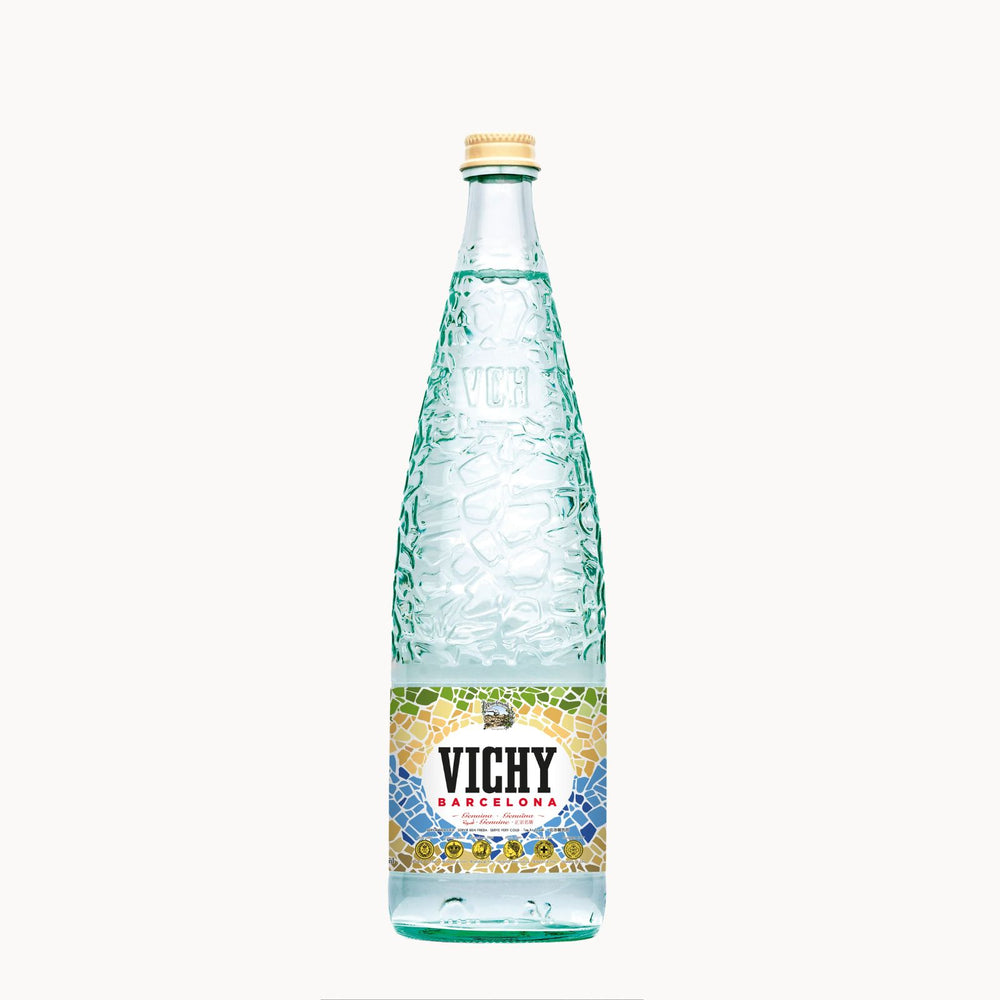 A glass bottle of Vichy Barcelona mineral water with a textured greenish-blue embossed pattern, a gold cap, and a colorful mosaic label, centered on a plain white background.