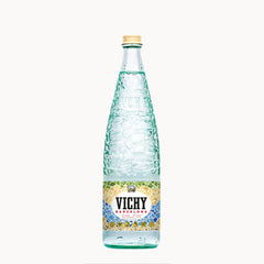 A glass bottle of Vichy Barcelona mineral water with a textured greenish-blue embossed pattern, a gold cap, and a colorful mosaic label, centered on a plain white background.