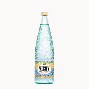 A glass bottle of Vichy Barcelona mineral water with a textured greenish-blue embossed pattern, a gold cap, and a colorful mosaic label, centered on a plain white background.