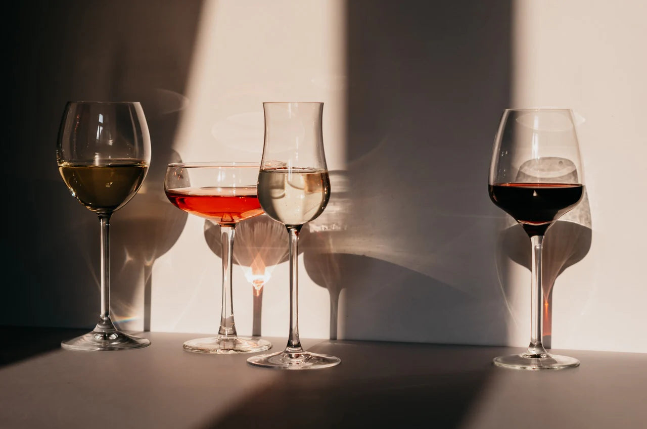 Four wine glasses with different types of wine on a dark surface with a light source creating shadows.