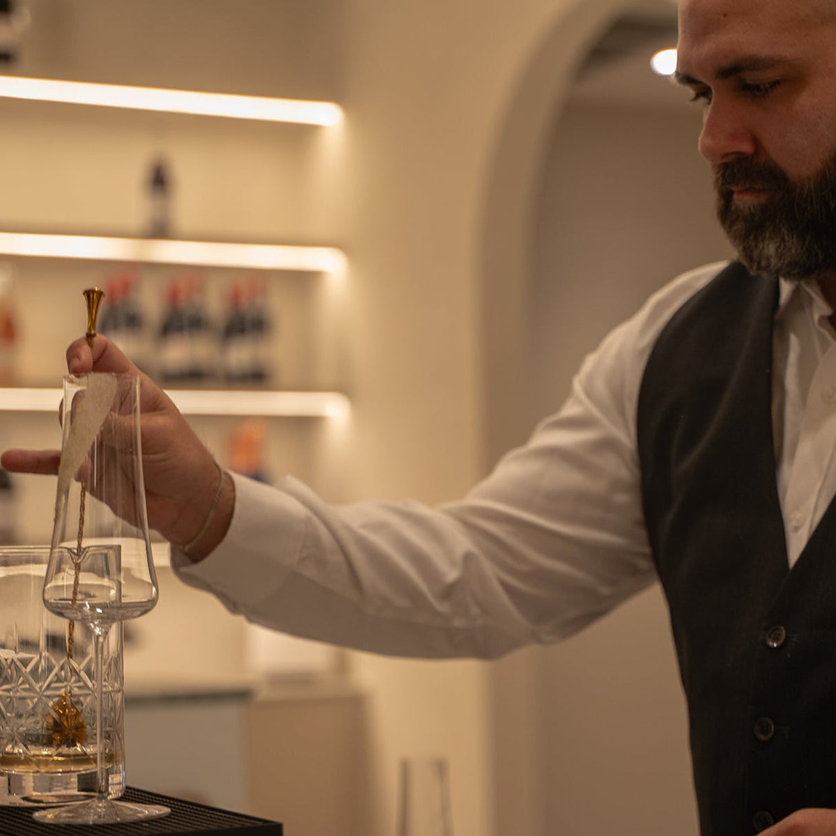 A bartender with a beard, wearing a white shirt and black vest, carefully pouring a clear liquid from a glass decanter into a glass, set in a warmly lit bar environment.